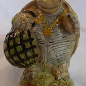 Puede incluir: Figura de tortuga de cerámica con un collar amarillo y una bolsa verde y negra. La tortuga es de color marrón y beige con un patrón de caparazón detallado. La bolsa se sostiene en la mano izquierda.