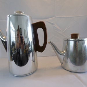 Puede incluir: Dos cafeteras de plata con asas de madera. La cafetera más grande tiene una superficie brillante y reflectante. La cafetera más pequeña tiene una superficie texturizada.