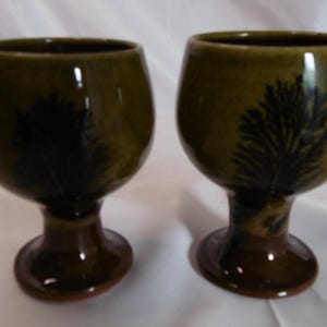 May include: Two ceramic wine goblets with a dark green glaze and a dark tree-like design. The stem and base are a darker brown. The goblets are on a white surface.