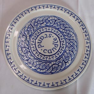 Puede incluir: Un plato de cerámica blanco con un diseño de nudo celta azul. El plato tiene un patrón de borde azul repetido y las palabras "candle.and.pubs" en el centro.