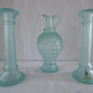 Op de afbeelding: Set van drie frosted glazen vazen in een lichtblauwe kleur. Twee zijn hoog en cilindrisch, terwijl de derde een kan is met een bolvormig lichaam en een getextureerd oppervlak.