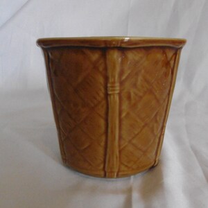 May include: A glossy, medium-brown ceramic planter featuring a raised, textured basket-weave pattern across its exterior surface. The pot has a wide, open top and vertical structural ribs that divide the woven design into distinct panels.