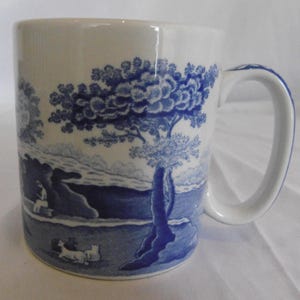 May include: White ceramic mug with a blue landscape design. The design features trees, figures, and a body of water. The handle is white with blue accents. The mug has a cylindrical shape.