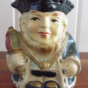 Puede incluir: Jarra de cerámica vintage con forma de hombre, que lleva un sombrero oscuro y una túnica verde y dorada. La jarra tiene una cara sonriente con labios rojos y sostiene una linterna. La jarra es un objeto decorativo.