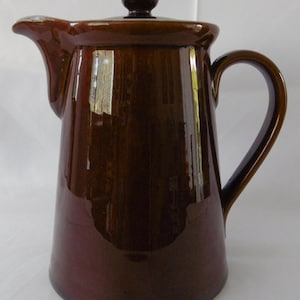 May include: A tall, brown ceramic coffee pot with a lid and handle. The pot has a glossy finish and a spout for pouring. The lid has a small, round knob. The handle is curved and attached to the side of the pot. The pot is likely used for serving hot beverages.