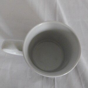 May include: A white ceramic mug with a curved handle. The interior of the mug is a light gray color. The mug is sitting on a white surface.