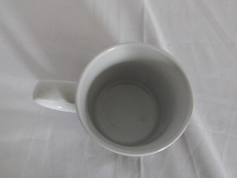 May include: A white ceramic mug with a curved handle. The interior of the mug is a light gray color. The mug is sitting on a white surface.
