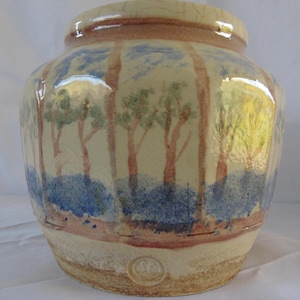 May include: A ceramic jar with a landscape design. The jar features a cream-colored base with a painted forest scene in shades of brown, blue, and green. The rim is a darker brown. The jar has a round shape and a wide opening.
