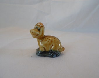 May include: A small ceramic figurine of a brown and tan dinosaur with a spotted pattern. The dinosaur is sitting on a blue and green base.
