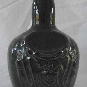 May include: A dark green ceramic bottle with a raised design of a knight on horseback. The bottle has a round body and a tall neck.