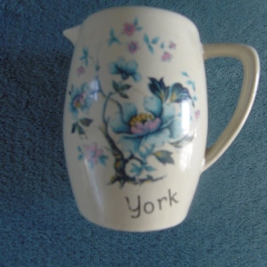 Może przedstawiać: Biały dzbanek ceramiczny z kwiatowym wzorem w odcieniach niebieskiego, różowego i zielonego. Dzbanek ma uchwyt, a na spodzie nadrukowano napis "York".
