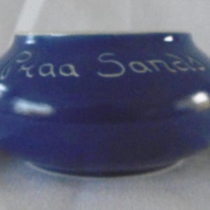 Puede incluir: Un pequeño cuenco redondo de cerámica azul con las palabras "Praa Sands" escritas en escritura blanca. El cuenco tiene una abertura ancha y un acabado liso y brillante. El texto está ligeramente borroso.