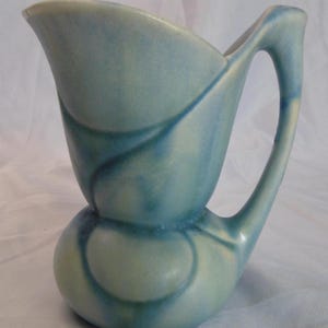 May include: A light blue ceramic jug with a distinctive, curved handle and a flared spout. The jug has a bulbous base and a design of swirling blue and white patterns. The overall height is approximately 18 cm.