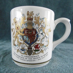 May include: White ceramic mug with a gold and colorful coat of arms featuring a lion and unicorn. The text reads "To commemorate the marriage of the Prince of Wales and Lady Diana Spencer - 29th July 1981."