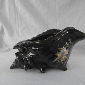 Peut inclure: Un pot de fleurs en céramique noire brillante en forme de coquillage. Le pot a un bord festonné et est orné d'un motif floral dans les tons de bleu, d'or et de vert. Il repose sur trois petits pieds.
