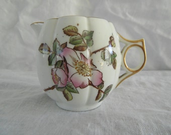 May include: A white porcelain creamer with a floral design. The creamer features a pink rose and green leaves, with gold trim on the handle and rim. The creamer has a scalloped edge and a small spout.