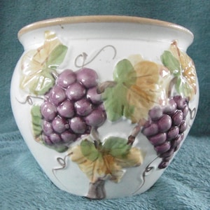 May include: White ceramic planter with a beige rim, featuring embossed purple grapes and green and yellow leaves. The planter has a rounded shape and is suitable for indoor or outdoor use.