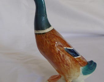 May include: A ceramic mallard duck figurine featuring a glossy finish with a dark green head, a white neck ring, a brown textured body, and orange webbed feet. The duck stands on a white base, with blue wing accents and an orange beak.
