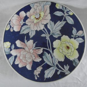 Puede incluir: Plato redondo de cerámica con fondo azul oscuro y diseño floral. Las flores son en tonos rosa y amarillo, con hojas verdes y grises. El plato tiene un borde blanco.