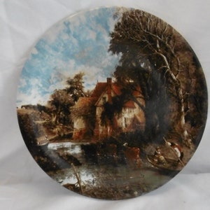 Puede incluir: Un plato redondo de cerámica con una imagen pintada de una escena campestre. La escena representa una casa de ladrillo rojo con un río en primer plano y árboles en el fondo. El cielo es azul con nubes blancas.