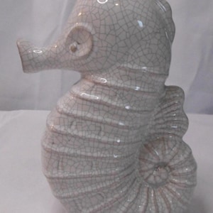 May include: A ceramic seahorse figurine in a crackle glaze finish. The seahorse is primarily white with subtle pink undertones and a textured surface. The tail is curled, and the figurine has a detailed, artistic design.