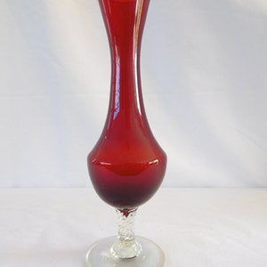 May include: A tall, red glass vase with a clear glass base. The vase has a slender neck and a rounded body. The base is twisted and has a flared foot.