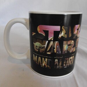 May include: White ceramic mug with a black background featuring the Star Wars: The Mandalorian logo and images of characters. The mug has a white handle and rim, perfect for coffee or tea.