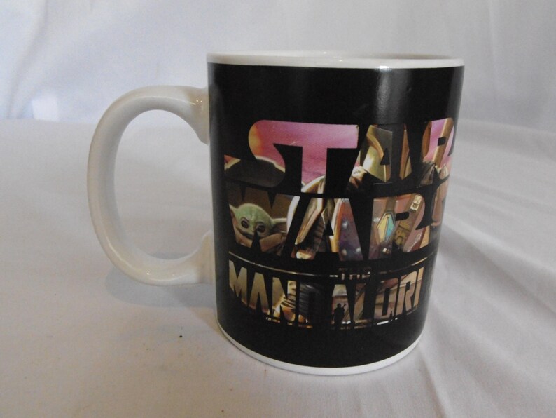 May include: White ceramic mug with a black background featuring the Star Wars: The Mandalorian logo and images of characters. The mug has a white handle and rim, perfect for coffee or tea.