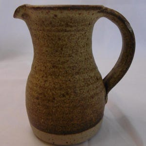 May include: A stoneware pitcher with a speckled tan and brown finish. The vessel features a curved handle, a flared rim with a pouring spout, and a rounded body that tapers toward the base, which has a lighter, unglazed band at the bottom edge.