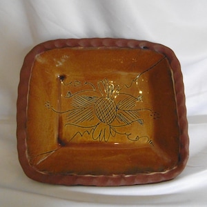 May include: A square ceramic dish with a glossy amber-coloured glaze and a dark brown scalloped rim. The centre of the dish features a hand-drawn black line art illustration of a stylised flower with radiating petals and thin, curved stems.