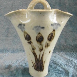 May include: Cream ceramic vase with a handle and a flared, scalloped rim. The vase has a hand-painted floral design in brown and blue. The vase is approximately 20 cm tall.