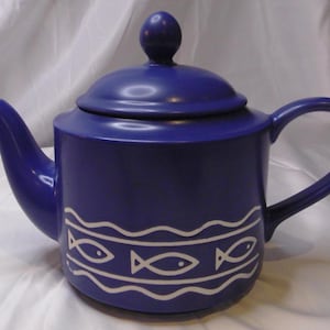 May include: A cobalt blue ceramic teapot with a matching lid and handle. The teapot features a white wave and fish design around the base. The spout and handle are curved. The teapot is approximately 7 inches tall.
