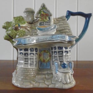 May include: A decorative ceramic teapot in the shape of a teahouse. The teapot has a blue handle, a grey roof, and a banner reading "Bluebirds Teahouse". The teahouse has a stone facade and a small wreath on the door.