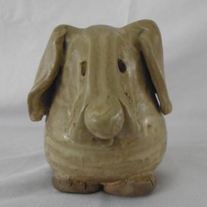 Peut inclure: Une sculpture en céramique d'un chien avec de grandes oreilles tombantes, un museau court et un corps rond. La sculpture est peinte dans un glacis brun clair.