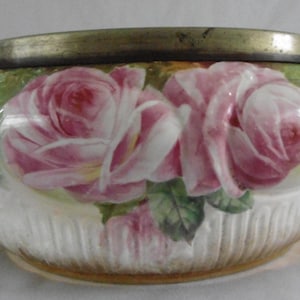 Könnte beinhalten: Dekorative Keramikschale mit Deckel, verziert mit rosa Rosen und grünen Blättern. Der Rand und der Deckel sind goldfarben. Der untere Teil der Schale hat eine gerippte Textur. Das Design erinnert an Vintage-Blumenmuster.