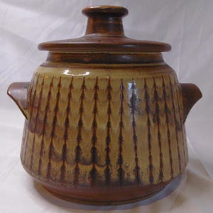 May include: A ceramic lidded pot with a textured, vertical pattern in shades of brown and tan. The pot has two side handles and a rounded base. The lid is flat with a small knob on top. The overall design is rustic and earthy.