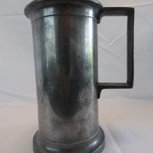 Puede incluir: Un jarro alto de peltre, cilíndrico, con un asa rectangular plana. La jarra tiene un borde ancho, ligeramente ensanchado y una base circular. El metal tiene un aspecto opaco y envejecido.
