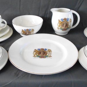 Op de afbeelding: Een witte porseleinen theeset met gouden rand, met een wapenontwerp. De set bevat kopjes, schotels, borden, een kom en een melkkan. Het wapenontwerp bevat een kroon, leeuwen en een portret. De set is op een donkere ondergrond geplaatst.
