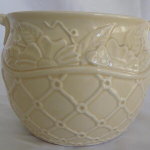 May include: Cream-coloured ceramic planter with a lattice pattern on the lower half and floral relief details around the top. The planter has two small handles and a rounded shape, suitable for indoor or outdoor use.