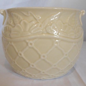 Puede incluir: Una maceta de cerámica color crema con un patrón de enrejado y relieve floral. La maceta tiene dos asas pequeñas y una forma redondeada. El diseño incluye un patrón floral detallado alrededor del borde.