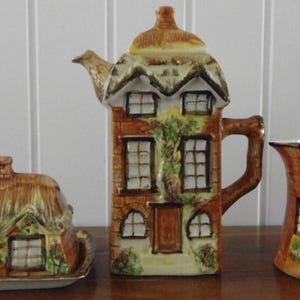May include: A ceramic tea set with a cottage design. The set includes a teapot, creamer jug, and a covered butter dish. The teapot is tall and rectangular, with a thatched roof and windows. The creamer and butter dish also have cottage designs.