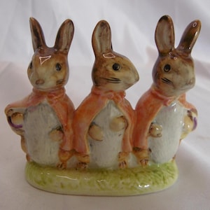 May include: A ceramic figurine featuring three brown rabbits wearing orange capes. The rabbits stand on a green base, with each holding a small basket. The figurine has a glossy finish and is likely a decorative item.
