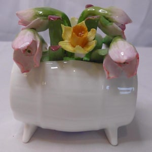Può includere: Un vaso di fiori in ceramica bianca con tre piccole gambe, riempito con una colorata composizione floreale. I fiori includono boccioli rosa e verdi e un narciso giallo. Il vaso ha una finitura lucida.