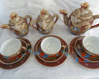 May include: A Japanese tea set with a dragon design. The set includes a teapot, creamer, sugar bowl, two teacups, and two saucers. The teacups and saucers have a red and gold floral design.