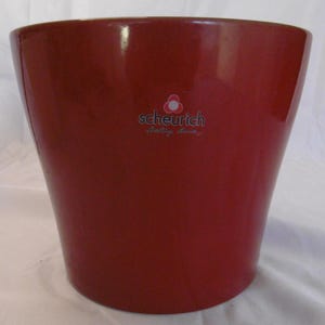 Può includere: Un vaso di ceramica rosso scuro lucido con una forma leggermente svasata. Il vaso ha una superficie liscia e un logo bianco con la scritta "scheurich".