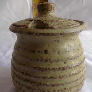 May include: A small, lidded ceramic jar with a light yellow base and brown speckled details. The jar has a rounded body with horizontal ridges and a matching lid. The jar is approximately 10 cm tall.