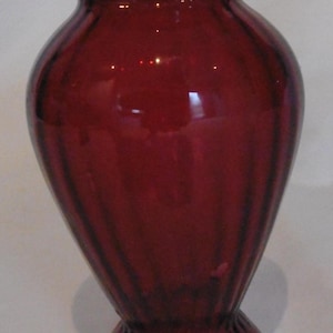 May include: A deep red glass vase featuring a flared rim, a rounded body, and a circular base. The entire surface of the vase is decorated with vertical ribbed detailing that extends from the base up to the neck, creating a textured appearance.