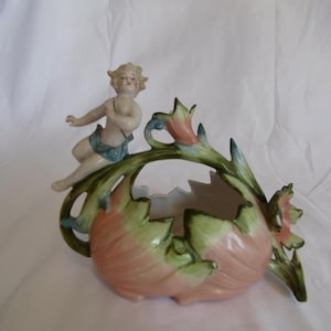 May include: A porcelain bowl shaped like a large, open pink and green flower with jagged edges. A small, pale-skinned figure with blonde hair and a blue cloth draped around the waist sits perched on the rim of the floral vessel.