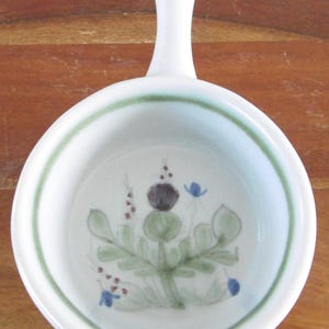 May include: White ceramic bowl with a handle, featuring a green border and a floral design with green leaves, a purple flower, and blue accents. The bowl is round and sits on a wooden surface.