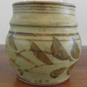 May include: A handcrafted ceramic jar with a pale green base and brown leaf and line patterns. The jar has a cylindrical form, wider at the base and narrower at the opening. The rim and upper section of the jar are a darker brown.